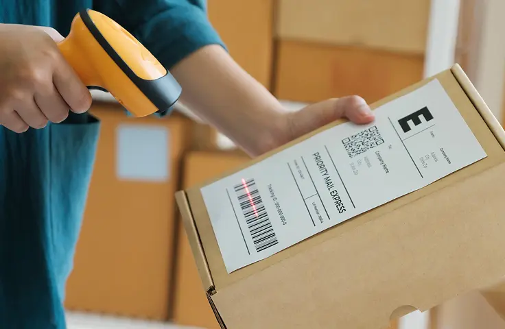 Picture this: a bustling warehouse where efficiency meets precision. Imagine the scene—a dedicated individual effortlessly scanning a barcode on a cardboard package with the ease of a maestro conducting an orchestra. This moment ensures seamless fulfillment, much like putting that final touch on your favorite recipe. Delicately perched on the front, you’ll find a shipping label adorned with both QR code and text—every detail meticulously in place, just as secure as Grandmas secret apple pie recipe. In the background, other packages wait patiently in neat stacks, their own journeys orchestrated to perfection. Now heres where ShipMonk.com comes into play—a true hero behind the scenes. Theyre like those old friends who always come through when you need them most; making sure every part of your logistics operation is as smooth as silk and every delivery warmly wrapped in reliability. Imagine Tom Hanks giving you his reassuring nod—that’s how ShipMonk makes you feel about your shipments!. via ShipMonk.com