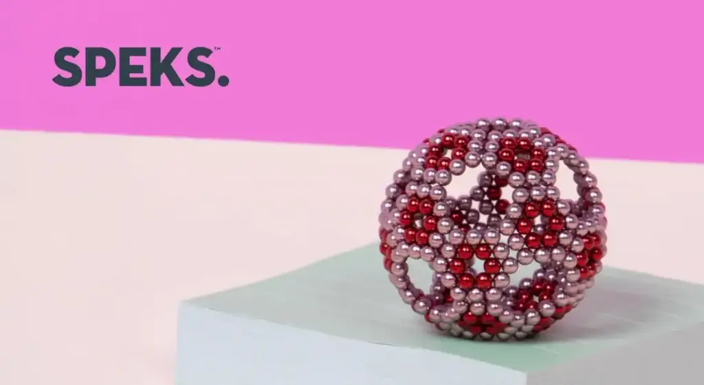 A geometric sphere made of red and silver beads sits on a light blue and white block against a pink background. The word SPEKS. is in the top left corner.