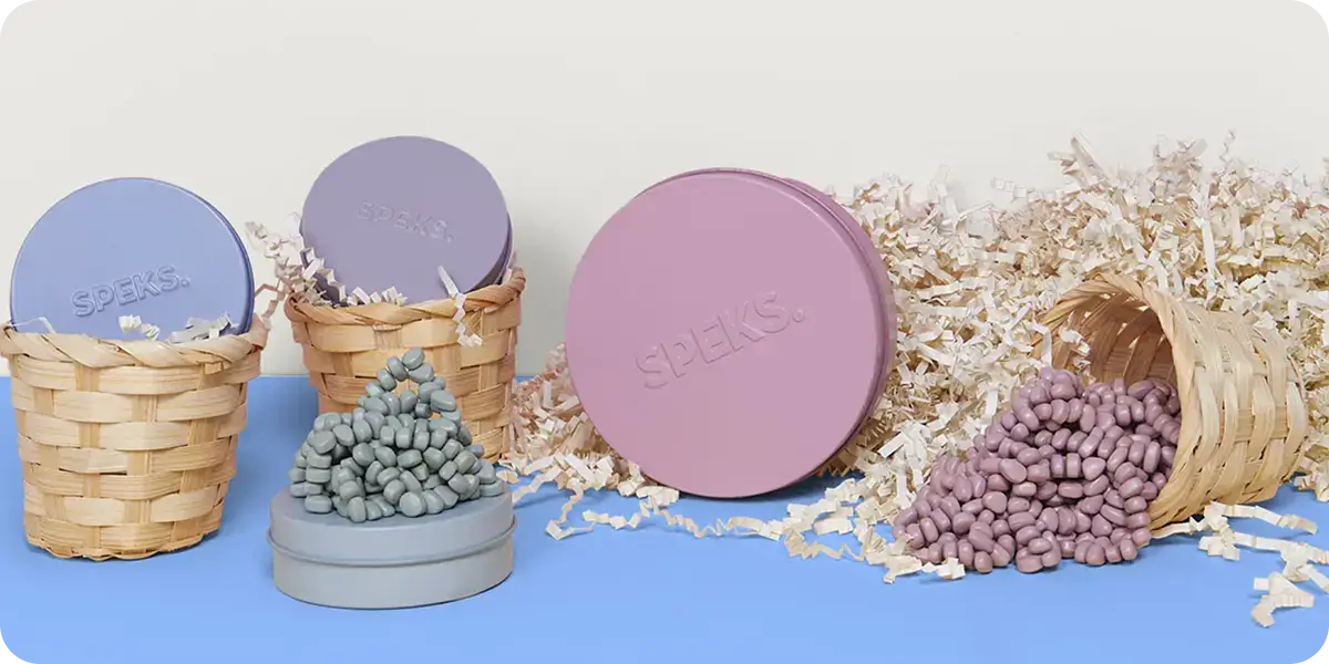 Round tins labeled SPEKS in pastel colors are displayed with small magnetic beads in matching shades. Beads are arranged in stacks and spilling from baskets amidst shredded paper, set on a blue and white background.