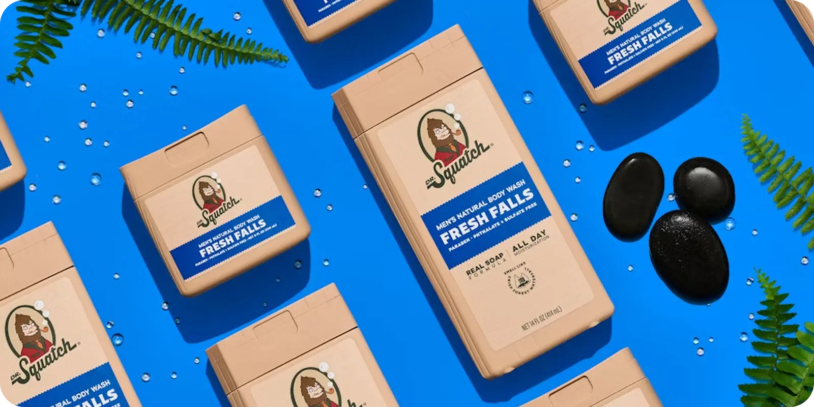 Imagine several Dr. Squatch Fresh Falls deodorant sticks, elegantly arranged on a lively blue background adorned with glistening water droplets, lush fern leaves, and sleek black stones—a scene that’s as inviting as it is refreshing. The tan packaging with its crisp blue label hints at nature’s best. Thanks to ShipMonk.com, this serene moment can arrive at your doorstep in a snap—bringing top-notch organization and a dose of everyday delight right to your routine. Isn’t that the kind of reliable magic we could all use?. via ShipMonk.com