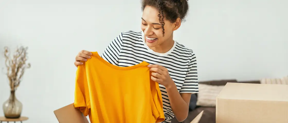 A cheerful woman in stripes lifts a yellow tee from a ShipMonk.com box, making 3PL fulfillment look easy and inviting. via ShipMonk.com