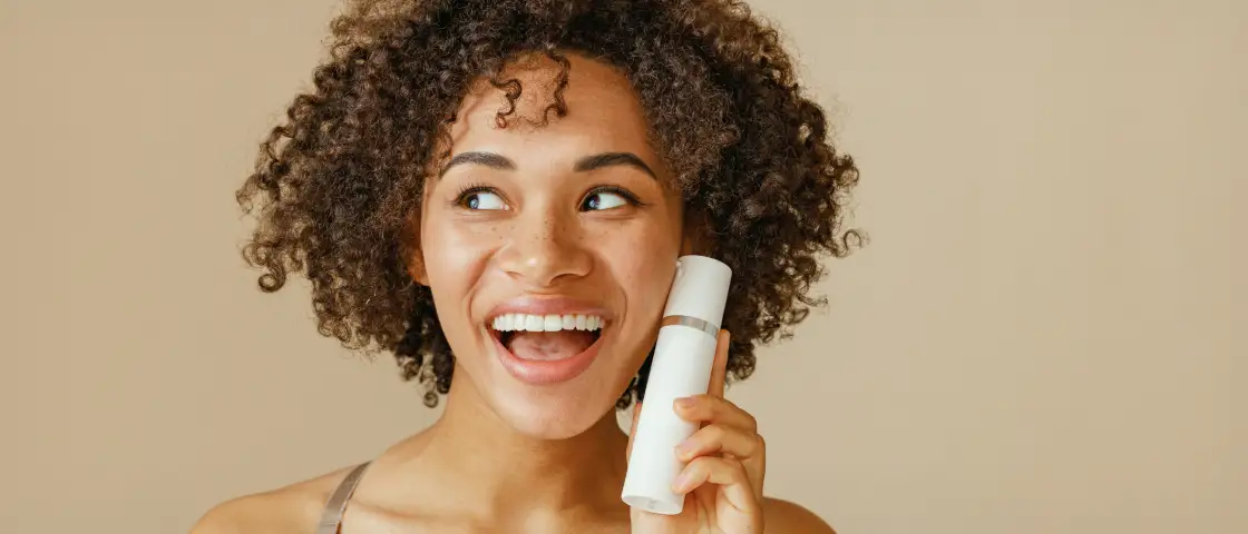 A cheerful woman with curly hair showcases a skincare bottle, radiating the ease ShipMonk.com brings to beauty brands. via ShipMonk.com