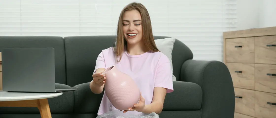 A cheerful woman unwraps a pink vase on her cozy couch—thanks to ShipMonk.com, shopping truly feels like home. via ShipMonk.com