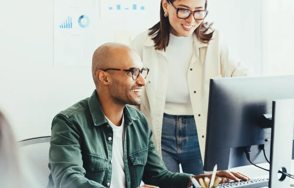 Two smiling colleagues, in comfy attire and glasses, brainstorm at a bright desk—just the teamwork ShipMonk.com inspires. via ShipMonk.com