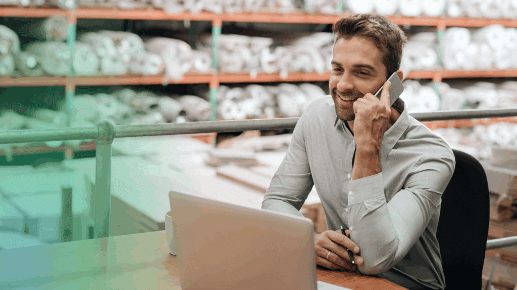 A cheerful man in a crisp shirt chats on the phone at his ShipMonk.com desk, laptop open and warehouse bustle behind him. via ShipMonk.com