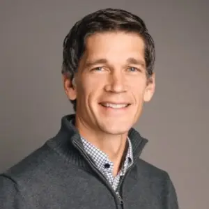 A friendly man with brown hair and blue eyes, in a gray sweater and checkered shirt, smiles warmly—ShipMonk style. via ShipMonk.com