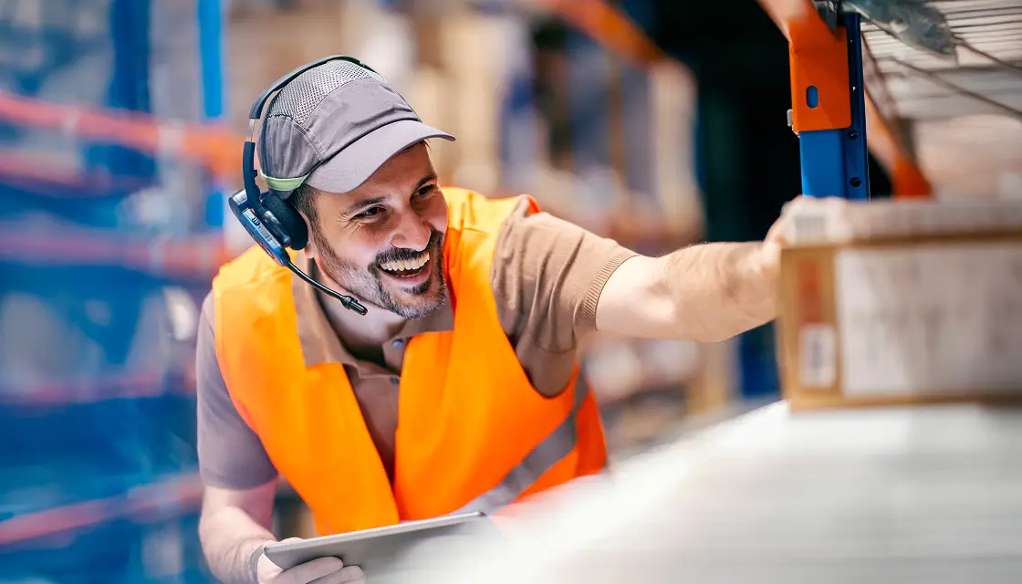 Smiling warmly, a ShipMonk.com team member in orange vest and cap checks inventory with tablet, ready to ship with care. via ShipMonk.com