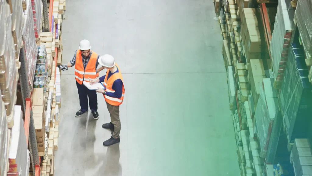 In a ShipMonk.com warehouse, two cheery workers in crisp orange vests and white helmets consult a clipboard amid towering boxes. via ShipMonk.com