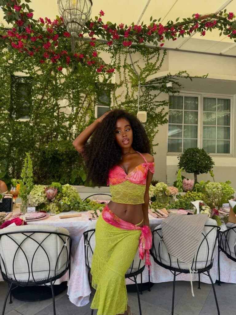A cheerful woman in a pink and yellow set grins by a lushly set outdoor table—wellness, shipped with care by ShipMonk.com. via ShipMonk.com
