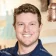 A cheerful man with wavy light brown hair and blue eyes beams in a navy jacket—classic ShipMonk.com warmth indoors. via ShipMonk.com